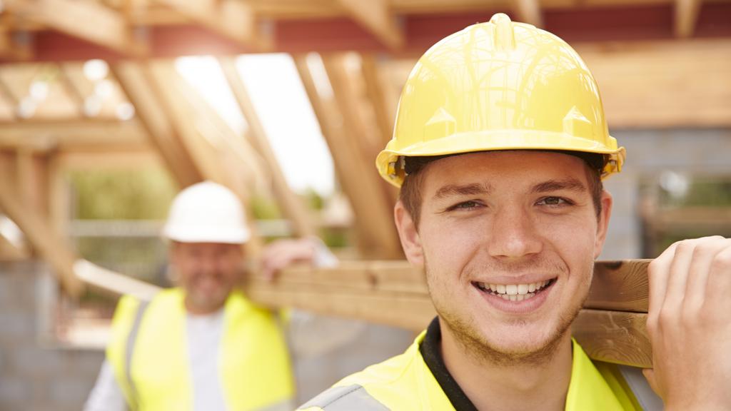 Builder And Apprentice Carrying Wood On Construction Site